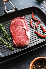 Wall Mural - Raw rib eye beef steak on black grilling pan with chili pepper and rosemary, top view.