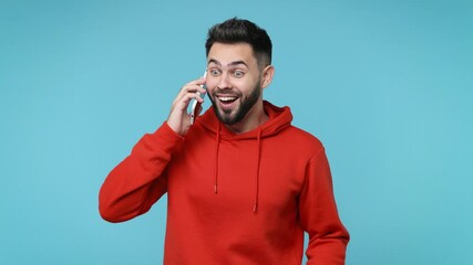 Wall Mural - Excited happy young bearded man 20s in casual red streetwear hoodie isolated on blue background studio. People lifestyle concept. Talk on mobile cell phone say yes doing winner gesture clenching fist
