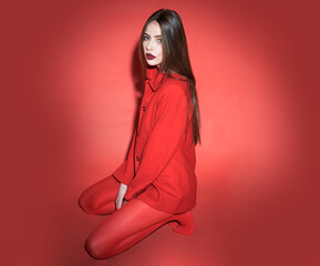 Wall Mural - Woman with posing in total red outfit. Fashion concept. Girl on calm face in red formal jacket and tights, red background. Lady looking at camera while sitting on floor.