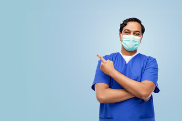 Wall Mural - healthcare, profession and medicine concept - indian doctor or male nurse in uniform and face protective medical mask showing something imaginary over blue background