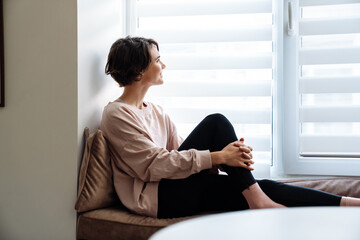 Happy beautiful girl smiling while sitting on windowsill at home