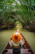 Picture from the back of a woman with an orange hat sitting on an old wooden ship. A Thai-style boat floats in a canal with many palm trees arching on either side. Feeling relax and wanderlust.