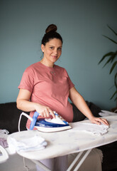 Wall Mural - Smiling pregnant woman at home ironing baby laundry.