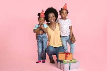 Poster - Portrait of young happy African mommy, hugging her two cute little kids daughters in party hats, celebrating birthday holiday party on pastel pink background with copy space