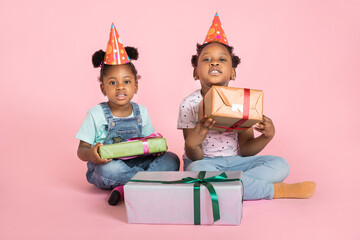 Poster - Two little adorable excited African girls, in red birthday hats, smiling and showing gift boxes to camera, sitting on the floor over pink pastel background. Birthday, holiday celebration concept.