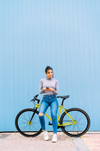 Woman Using Mobile Phone Looking Away While Sitting On Fixie Bike Against Blue Wall