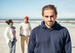 © Uwe Umst√§tter/Westend61 - Portrait of young man wearing hooded shirt standing on beach with two young woman walking behind in background