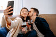 © Jake Jakab/ADDICTIVE STOCK - Content couple sitting on sofa with wineglasses and taking self portrait on smartphone while relaxing at weekend at home