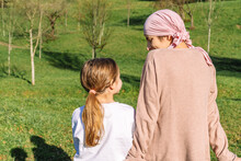 Back View Of Tender Calm Mother With Cancer Wearing Pink Head Scarf Speaking With Little Daughter Sitting On A Bench On Green Park
