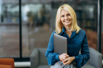 Portrait friendly elegant successful lady is looking at the camera,holding laptop in arms. Middle aged business woman in stylish wear stands in modern office and smiles