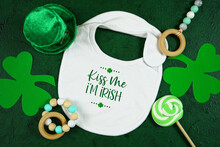 Happy St Patrick's Day Baby Wear White Bib, Styled With Leprechaun Hat, Shamrocks, On A Textured Green Background. Kiss Me I'm Irish Text.