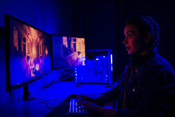 Wall Mural - A girl with headphones plays a video game on a computer on large monitors. Gamer with mouse and keyboard. Online games with friends, win.e-sports, streams.Teenagers play adventure games.Neon lighting