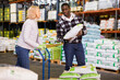 © JackF - Confident African American warehouse cheerful glad worker with mature female assistant loading sacks with fertilizers on trolley cart