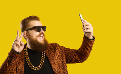 Eccentric rich man live video streaming on mobile phone. Happy confident guy in trendy glasses, rapper gold neck chain and leopard print suit jacket doing V sign and taking funny selfie on smartphone