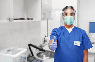 medicine, healthcare and protection concept - indian doctor or male nurse in blue uniform, protective medical mask and face shield showing thumbs up over laboratory or hospital background