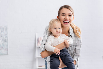 astonished woman looking at camera while holding smiling kid at home