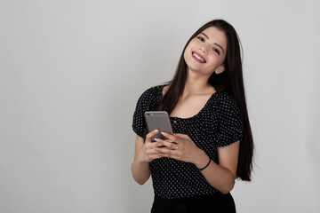 Portrait of pretty young Indian girl standing against white background and holding phone, sending message, video conference with friend, taking selfie.