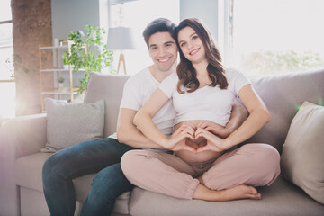 Poster - Photo of cheerful funny positive family waiting baby sitting sofa hugging tummy showing fingers heart closed eyes indoors inside