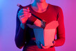 © Bisual Photo - Anonymous woman holding Asian noodles box for lunch with chopsticks in studio with blue and red lights