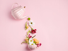 Creative Layout With Tea Pot Pouring Fresh Flowers And Leaves Into Tea Cup On Pastel Pink Background. Creative Floral Spring Bloom Concept. Still Life Visual Trend. Flat Lay.