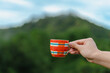 Close-up of a hand holding a colorful coffee cup with handle in the morning. Blurred background of lush mountains and trees. Feeling fresh and relax. Idea for lifestyle wallpaper with copy space.