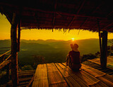 Fototapeta Zachód słońca - Picture from the back of a woman sitting on wooden porch extending into a high mountain cliff. The sun is setting on the mountain and there is a beautiful warm orange light. The traveling background.