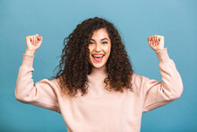 Photo Of Cheerful Beautiful Young Woman Standing Isolated Over Blue Wall Background. Looking Camera Showing Winner Gesture.
