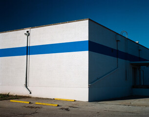 A minimal urban detail geometric study of the corner of a brilliantly white building with a blue stripe adorned with drain spouts, an access ladder and yellow parking curbs in the foreground.