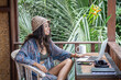© Lila Koan - Portrait of young brunette woman freelancer in a straw hat working with laptop on balcony of tropical bungalow with palm trees view.