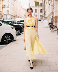 Street fashion. Beautiful young model lady walking on city road. Fashionable feminine look. Long yellow summer evening dress. Sleeveless dress with long pleating skirt.