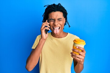 Wall Mural - Young african american man using smartphone and drinking a cup of coffee sticking tongue out happy with funny expression.