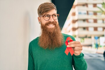 Sticker - Young irish man with redhead beard smiling happy and holding red hiv ribbon leaning on the wall at the city.