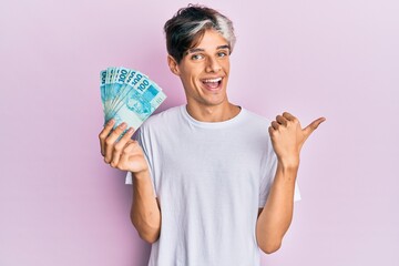 Young hispanic man holding 100 brazilian real banknotes pointing thumb up to the side smiling happy with open mouth