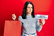 © Krakenimages.com - Middle age brunette woman holding merry christmas message and shopping bag puffing cheeks with funny face. mouth inflated with air, catching air.