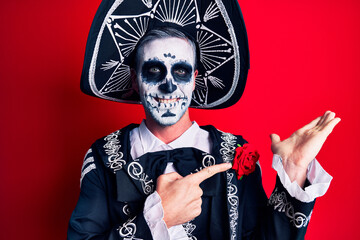 Wall Mural - Young man wearing mexican day of the dead costume over red amazed and smiling to the camera while presenting with hand and pointing with finger.