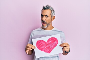 Poster - Middle age grey-haired man holding heart draw in shock face, looking skeptical and sarcastic, surprised with open mouth