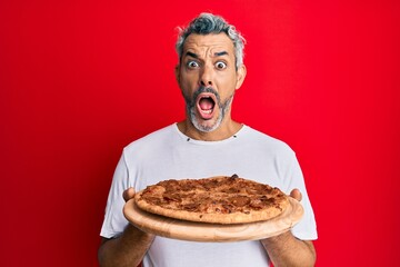 Poster - Middle age grey-haired man holding italian pizza afraid and shocked with surprise and amazed expression, fear and excited face.