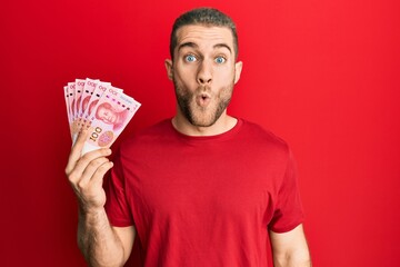 Wall Mural - Young caucasian man holding yuan chinese banknotes scared and amazed with open mouth for surprise, disbelief face