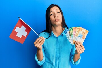 Poster - Young latin woman holding switzerland flag and franc banknotes depressed and worry for distress, crying angry and afraid. sad expression.