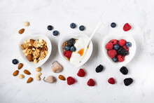 Trio Of Heart-shaped Bowls Arranged On A Slightly Textured Background With Berries, Dried Fruit, White Yogurt, Wholemeal Flakes And Honey