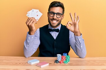 Sticker - Handsome hispanic croupier man sitting on the table with poker chips and cards doing ok sign with fingers, smiling friendly gesturing excellent symbol