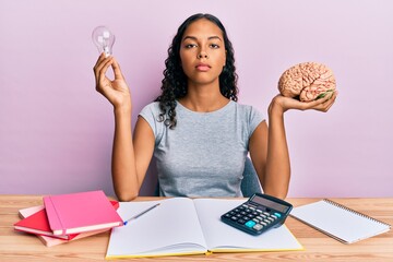 Wall Mural - Young african american girl holding brain and light bulb studying for school relaxed with serious expression on face. simple and natural looking at the camera.