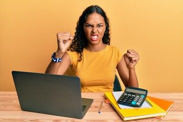 Sticker - Young african american girl working at the office with laptop and calculator angry and mad raising fists frustrated and furious while shouting with anger. rage and aggressive concept.
