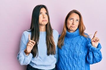 Wall Mural - Hispanic family of mother and daughter wearing wool winter sweater pointing up looking sad and upset, indicating direction with fingers, unhappy and depressed.