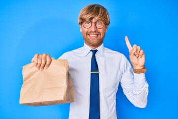 Poster - Young blond man wearing business clothes holding take away paper bag smiling with an idea or question pointing finger with happy face, number one