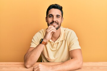 Young hispanic man wearing casual clothes sitting on the table looking confident at the camera with smile with crossed arms and hand raised on chin. thinking positive.