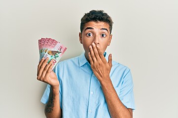 Sticker - Young handsome african american man holding 100 new zealand dollars banknote covering mouth with hand, shocked and afraid for mistake. surprised expression