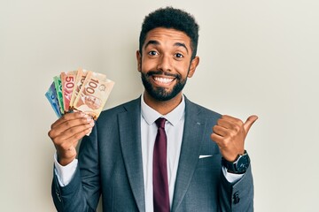 Canvas Print - Handsome hispanic business man with beard holding canadian dollars pointing thumb up to the side smiling happy with open mouth