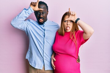 Canvas Print - Young interracial couple expecting a baby, touching pregnant belly making fun of people with fingers on forehead doing loser gesture mocking and insulting.