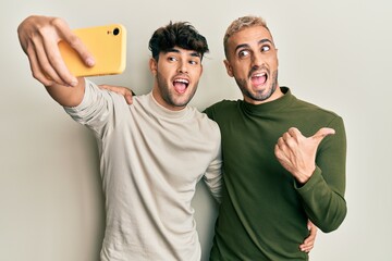 Homosexual gay couple standing together taking a selfie photo with smartphone pointing thumb up to the side smiling happy with open mouth